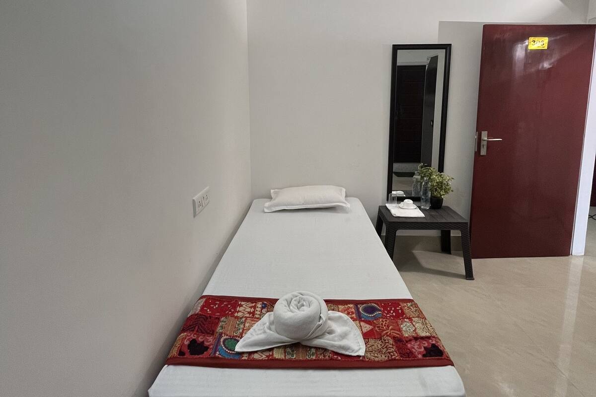 A single bed with a white mattress and a red patterned blanket, a small bedside table with a plant, a mirror, and a door with a yellow tag.