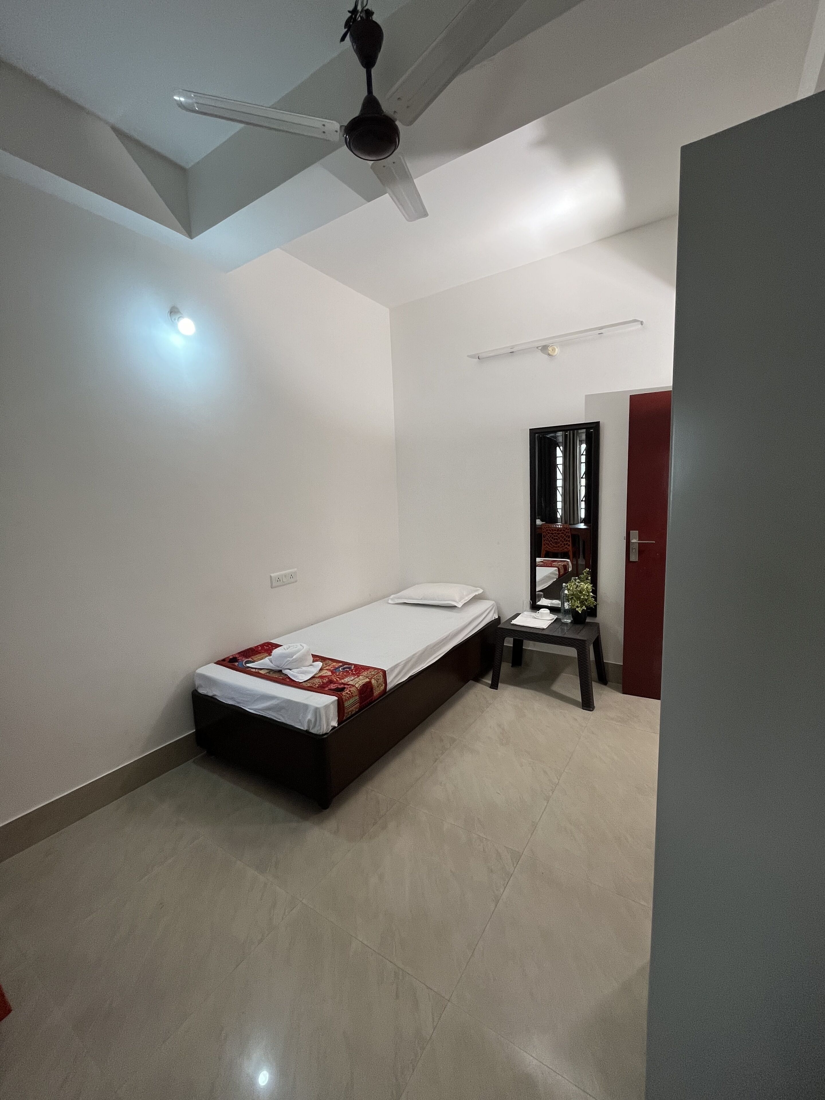 A single bed with a white mattress and a red blanket, a small table with a vase, a ceiling fan, and a red door.