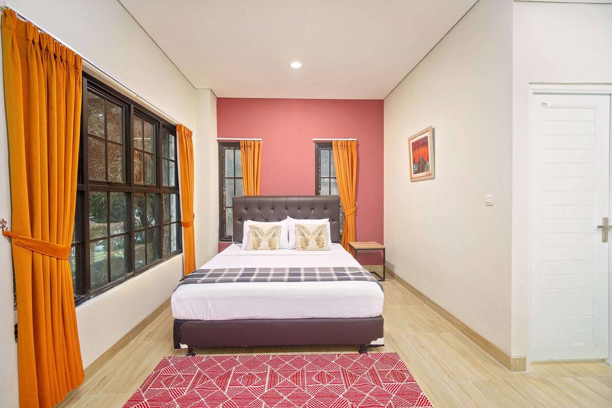 A bedroom with a bed, two windows with curtains, a nightstand, a door, and a wall-mounted picture.