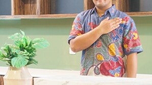 A man standing at a reception desk with a patterned shirt, a plant, and wooden shelves in the background. - Habitat at Gandaria (Jakarta)