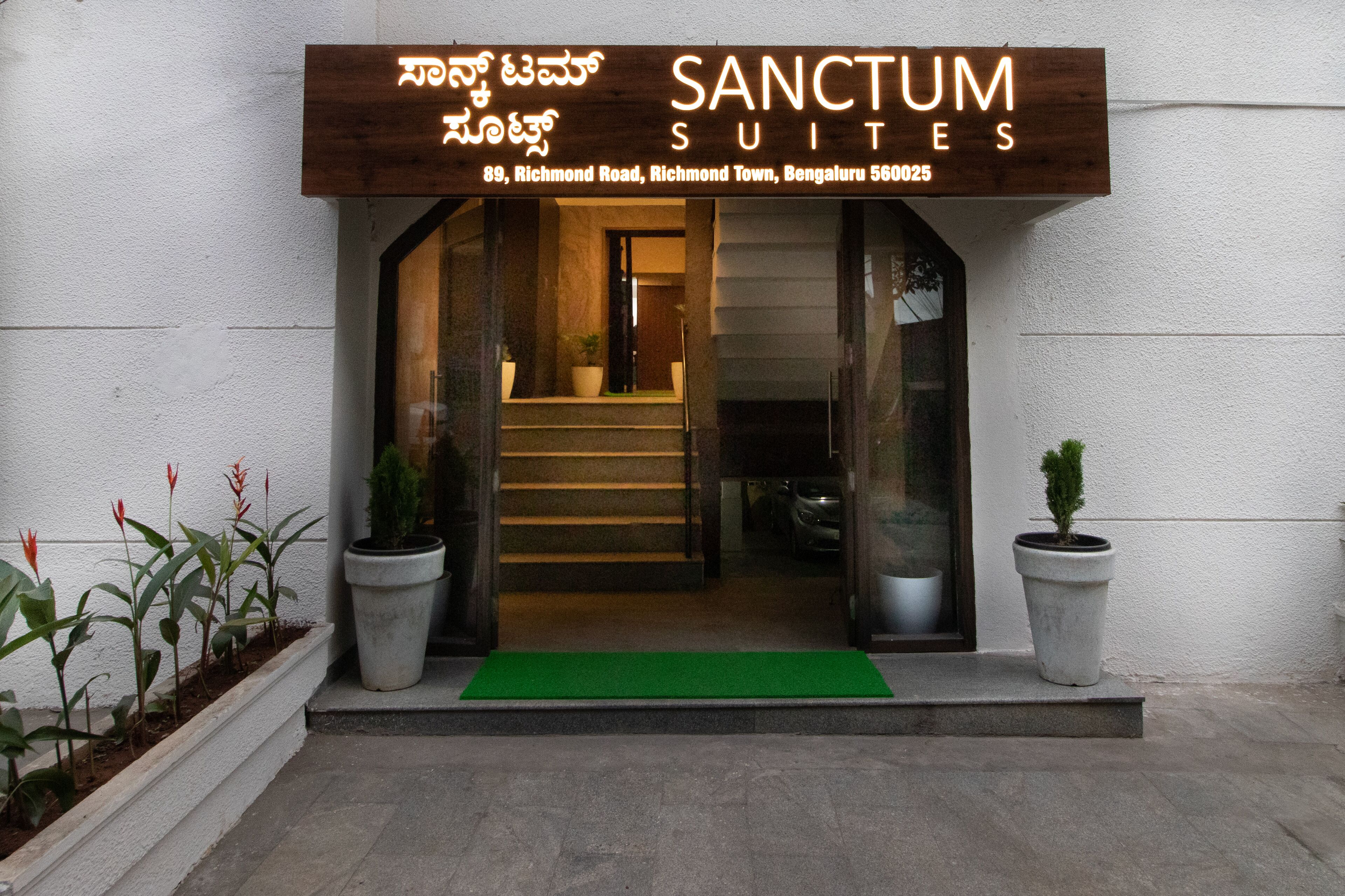The entrance to Sanctum Suites, featuring a wooden signboard with the name and address, a green mat at the entrance, and potted plants on either side.