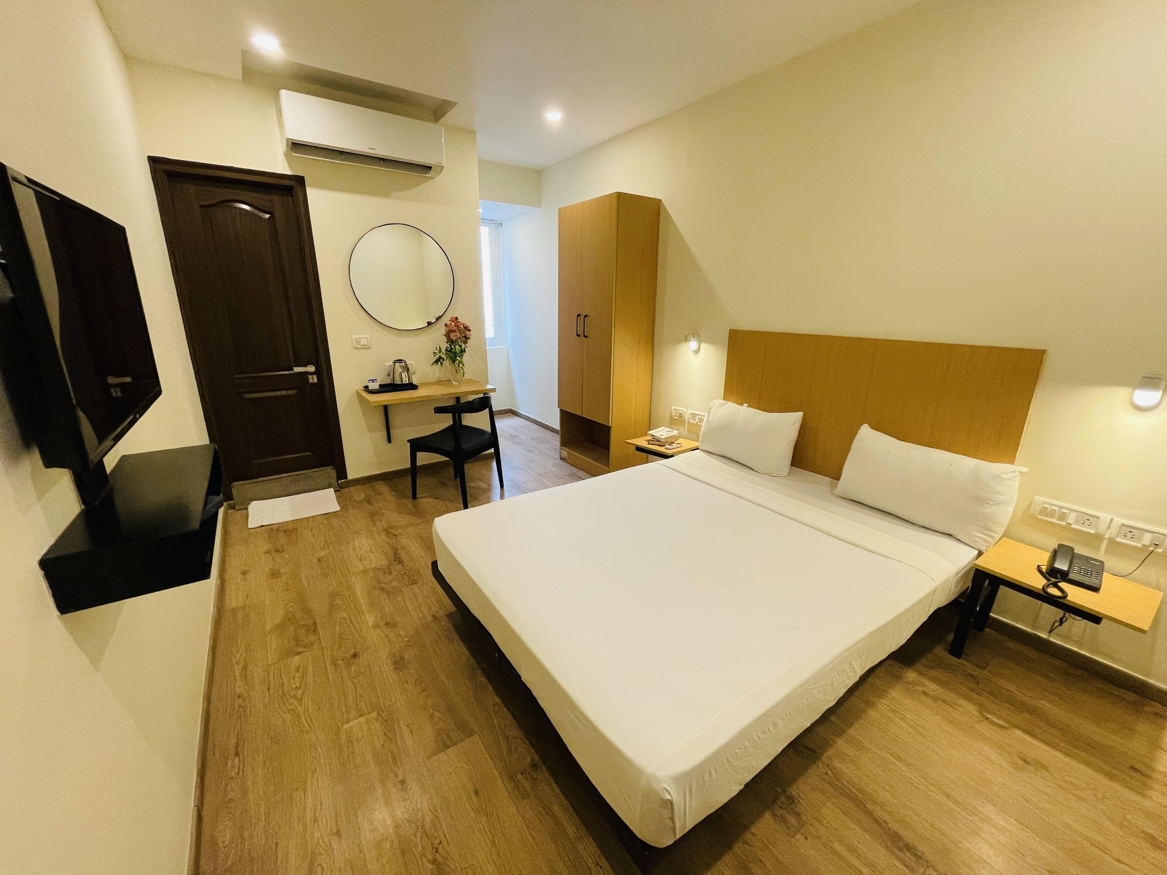 A hotel room with a large bed, a desk with a chair, a TV, a mirror, and a wooden wardrobe.
