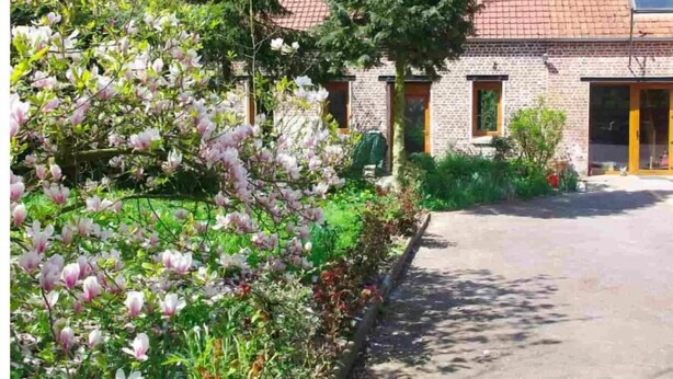 Un edificio de ladrillo con techo de tejas, camino de entrada pavimentado y jardín con árboles floreados.