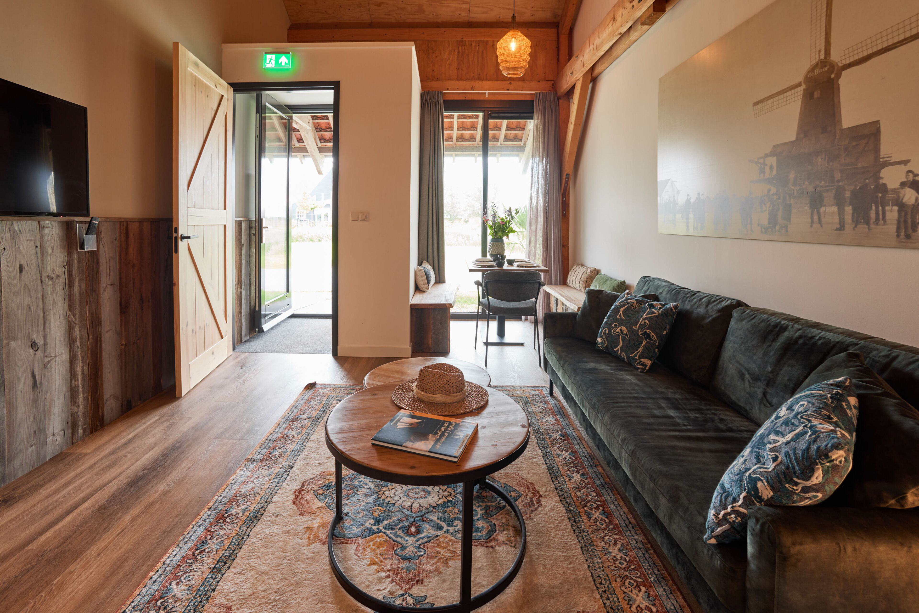 A cozy room with a wooden floor, a dark green sofa, a round coffee table with a hat and book, and a large window with curtains.