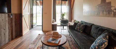 A cozy room with a wooden floor, a dark green sofa, a round coffee table with a hat and book, and a large window with curtains.