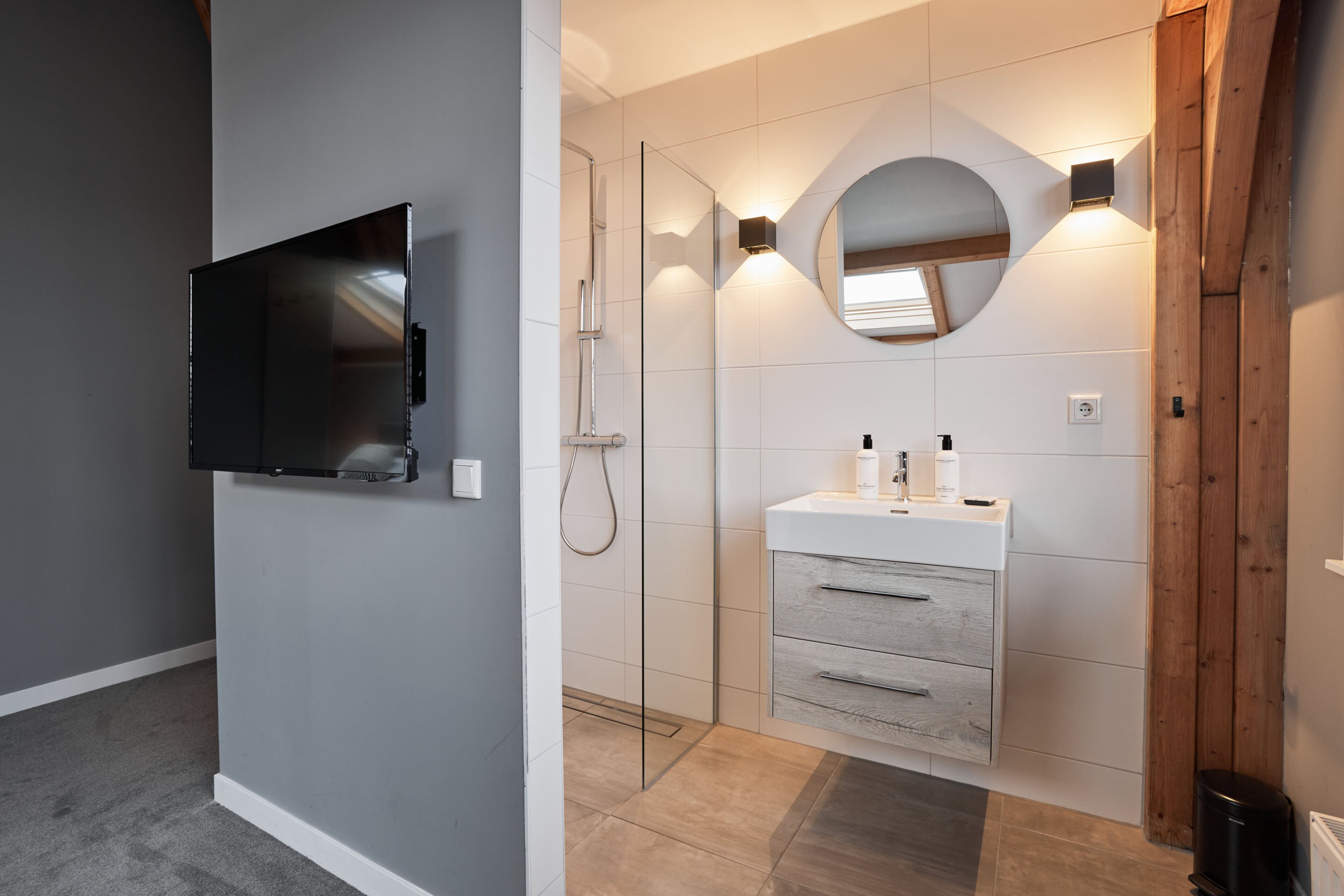 Une salle de bain moderne dotée d’un téléviseur encastré, d’une douche en verre, d’un meuble-lavabo avec miroir et d’un mur d’accent en bois.