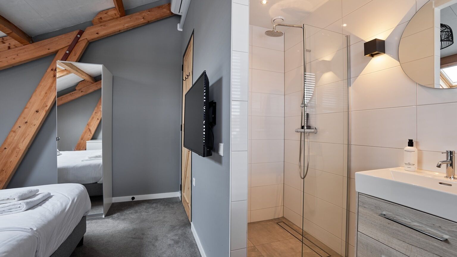 A modern hotel room with a sloped ceiling, a shower with a glass door, a mounted TV, and a white vanity with a mirror.