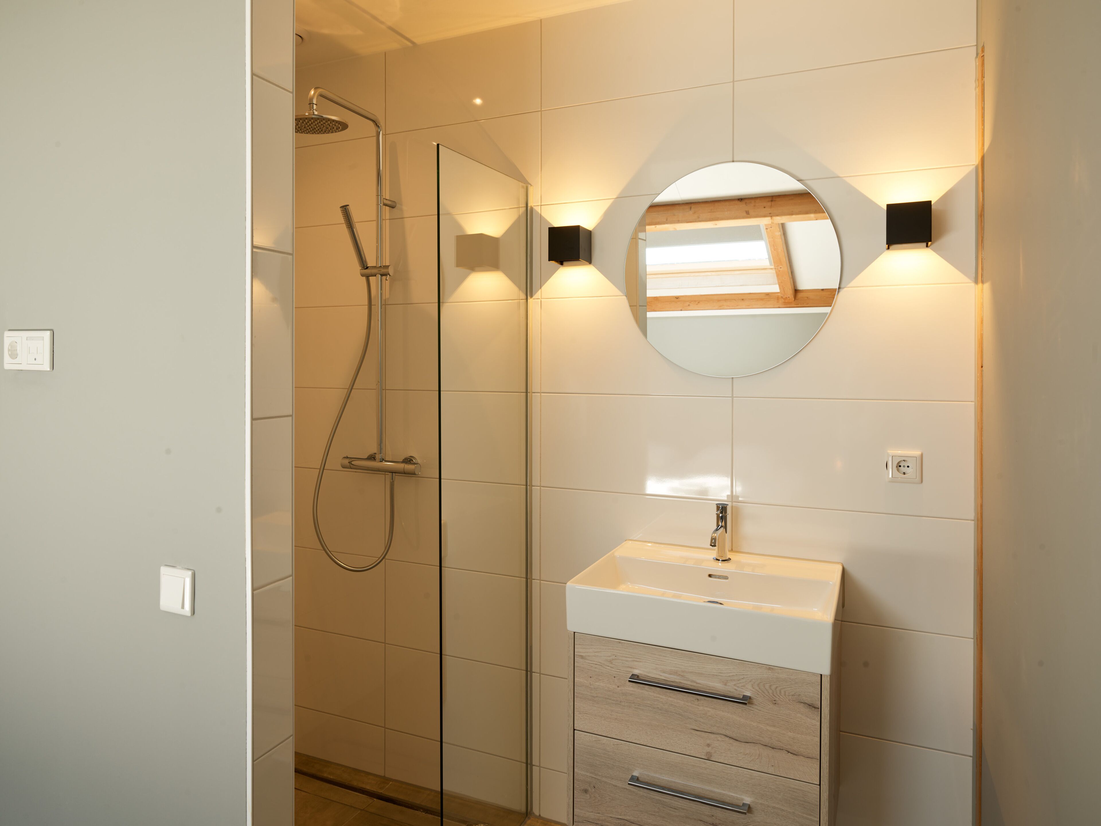 Een moderne badkamer met een douchecabine van glas, een ronde spiegel en een wit wastafelmeubel met houten lades.