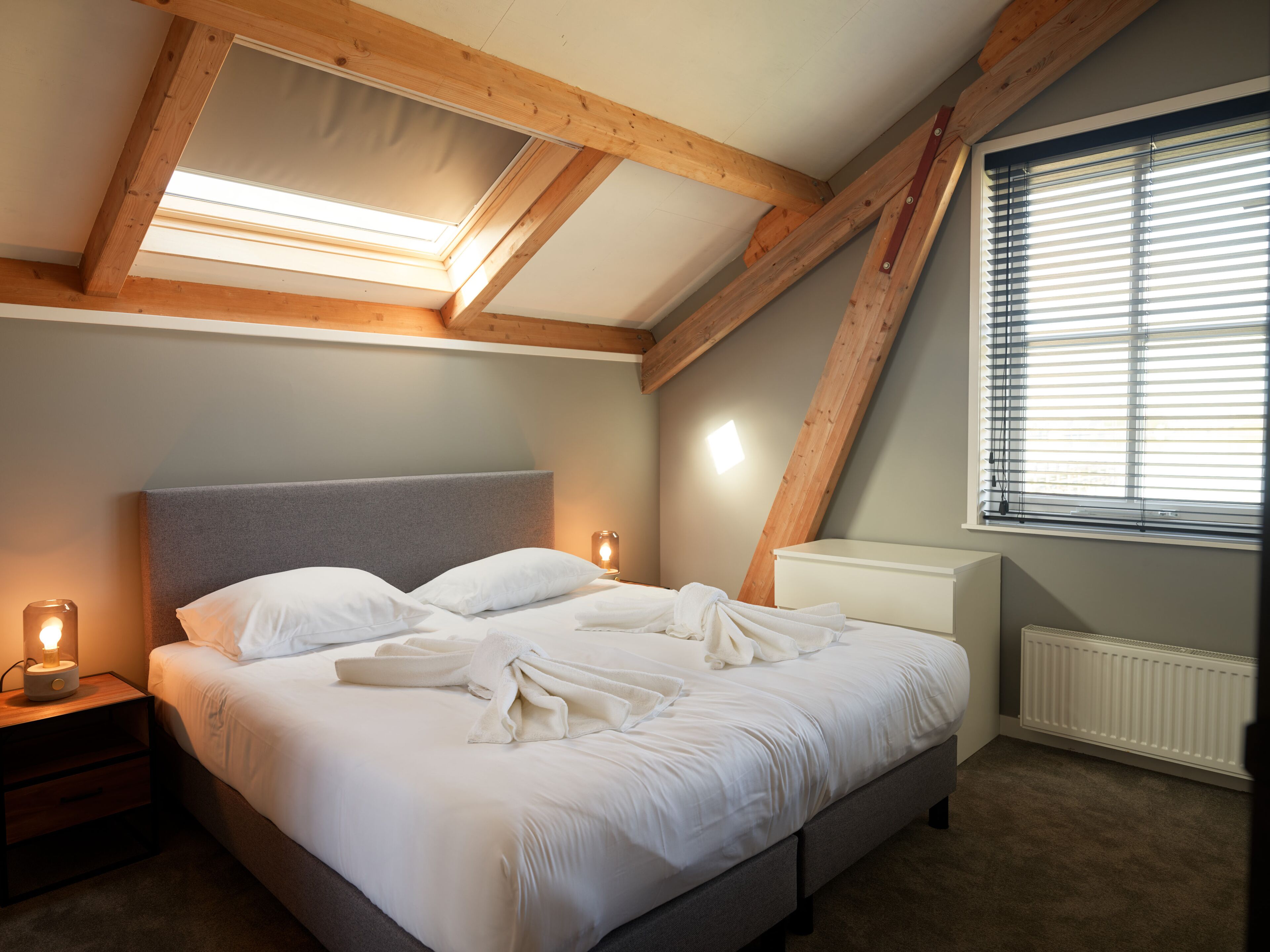 Een slaapkamer met een schuine balkenlaag, een bed met witte lakens, een nachtkastje met een lamp en een raam met jaloezieën.