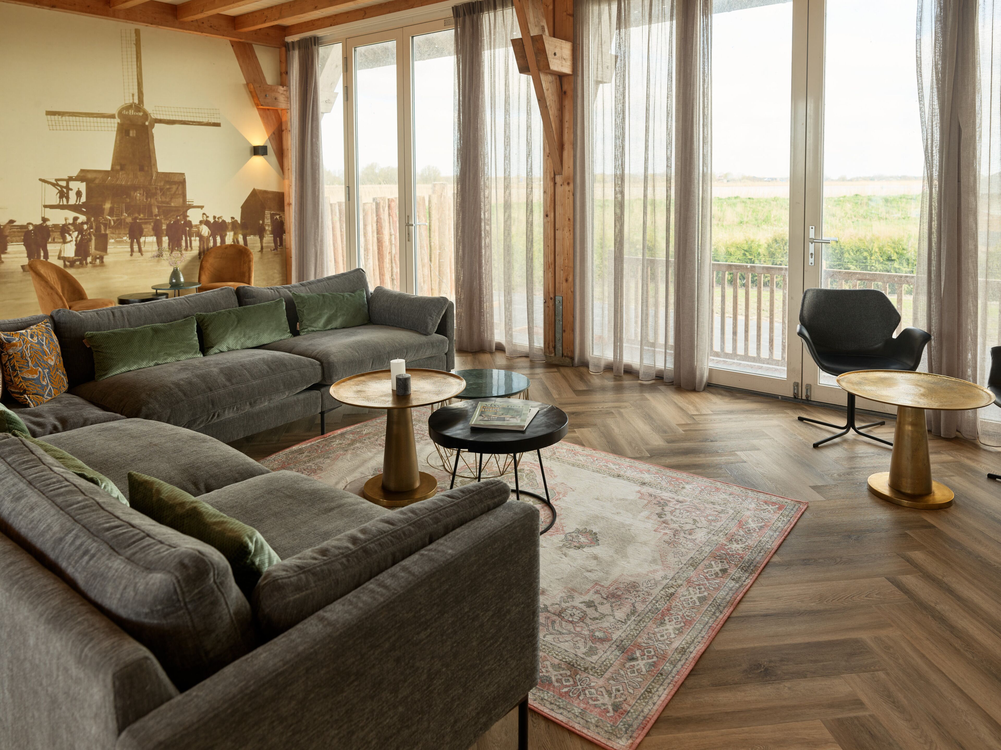 A spacious living area with a large sectional sofa, a round coffee table, and a black armchair. A large window with sheer curtains offers a view of the outdoors.