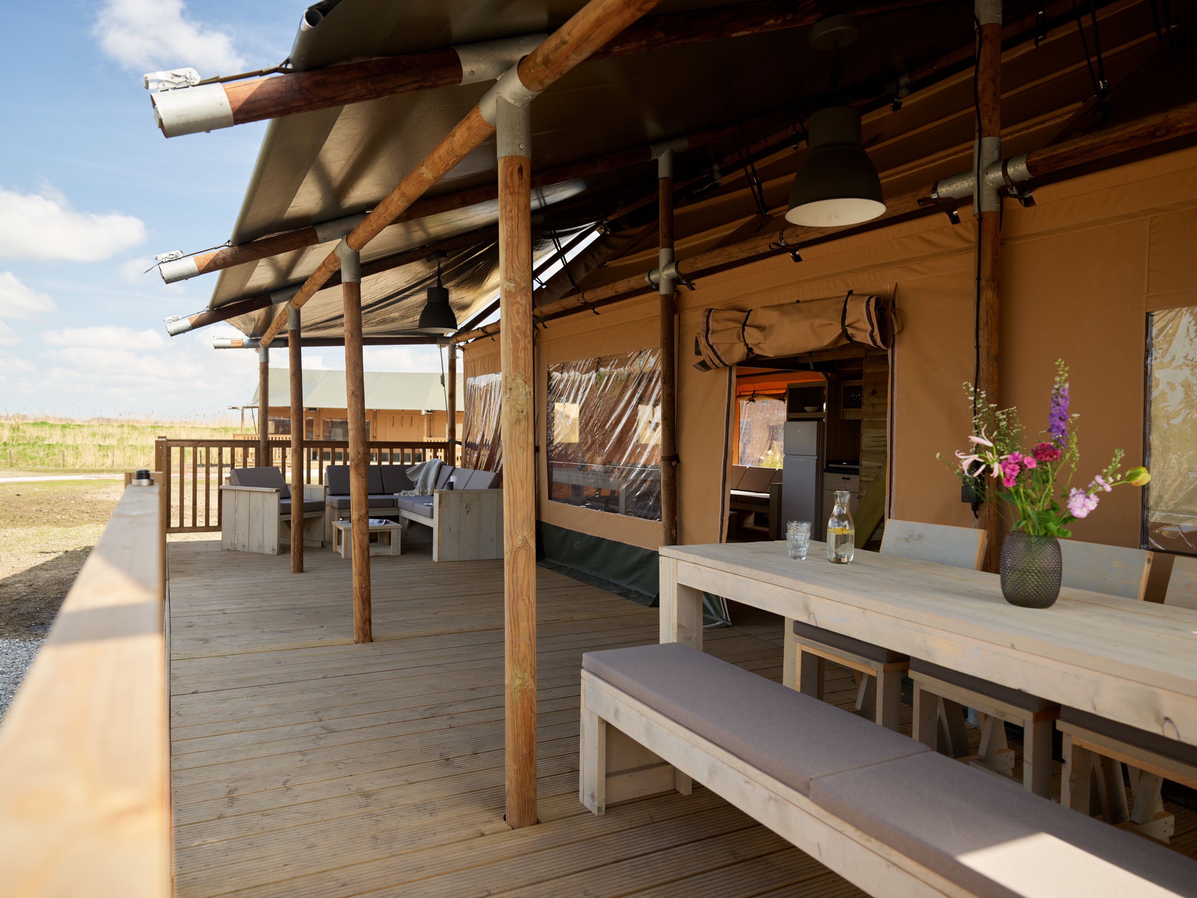 Une terrasse extérieure en bois, dotée d’un espace salon couvert, d’une table ornée d’un vase de fleurs et offrant une vue sur le paysage environnant.