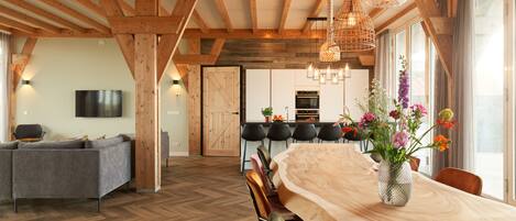 A spacious living area with a wooden ceiling, a dining table with chairs, a sofa, and a TV mounted on the wall.