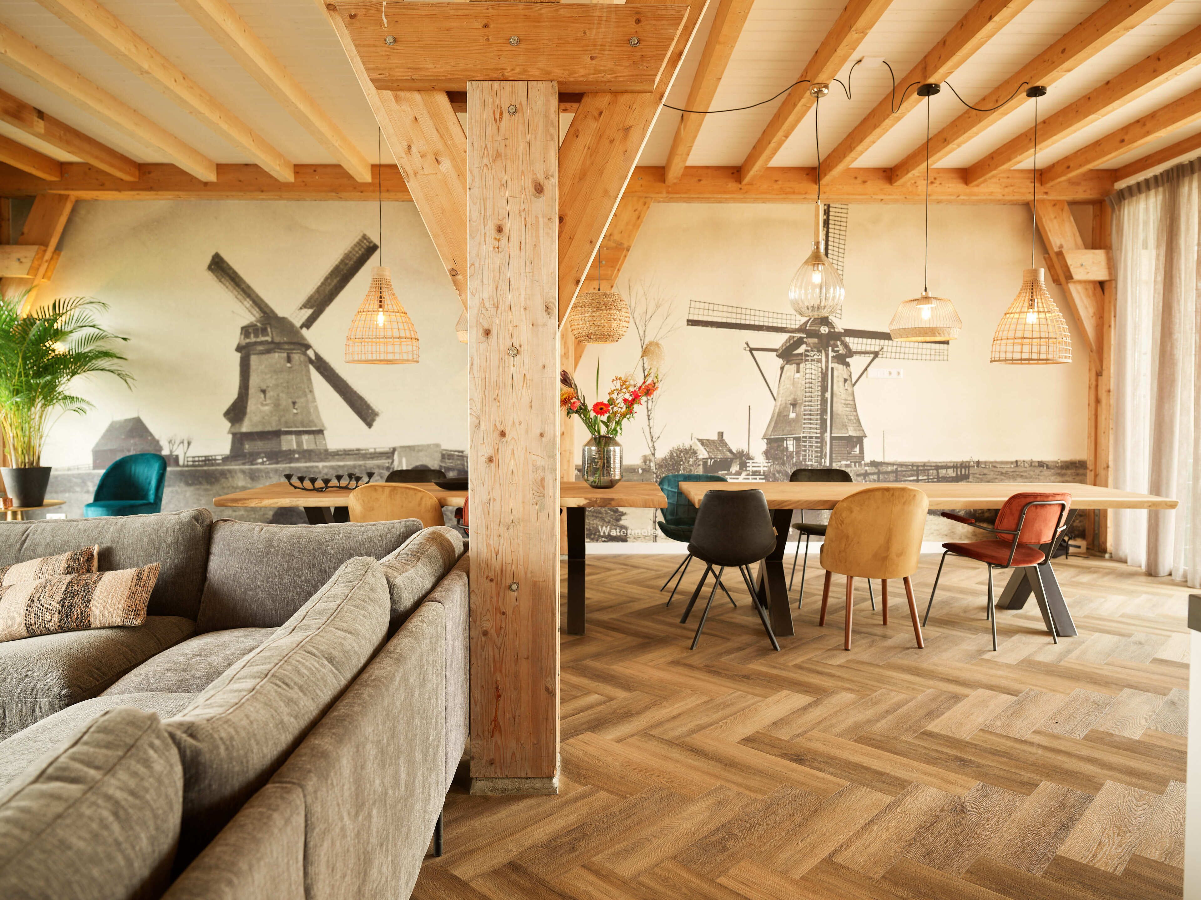 A spacious room with a herringbone wooden floor, a wooden dining table with chairs, a sofa, and a large window with a windmill mural.