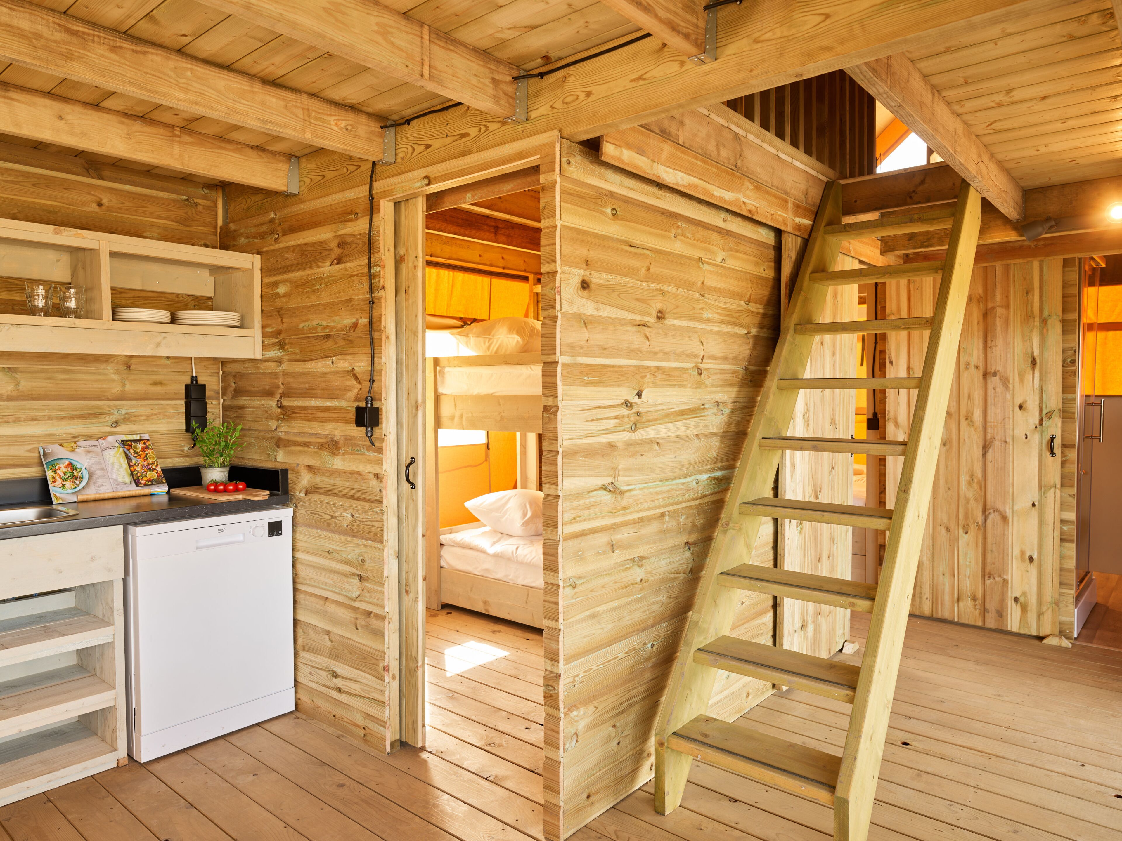 A wooden cabin interior with a kitchen area, a sleeping area, and a wooden ladder.