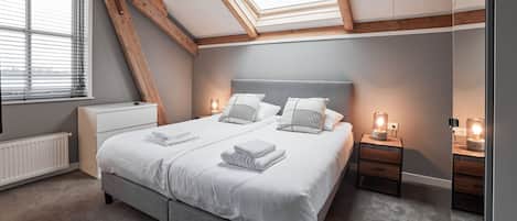 Un dormitorio con techo estilo loft, una cama con ropa de cama blanca, dos mesitas de noche con lámparas y un radiador.