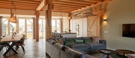 A spacious living area with a large sectional sofa, wooden beams on the ceiling, a dining table with chairs, and a kitchen area with a large window.