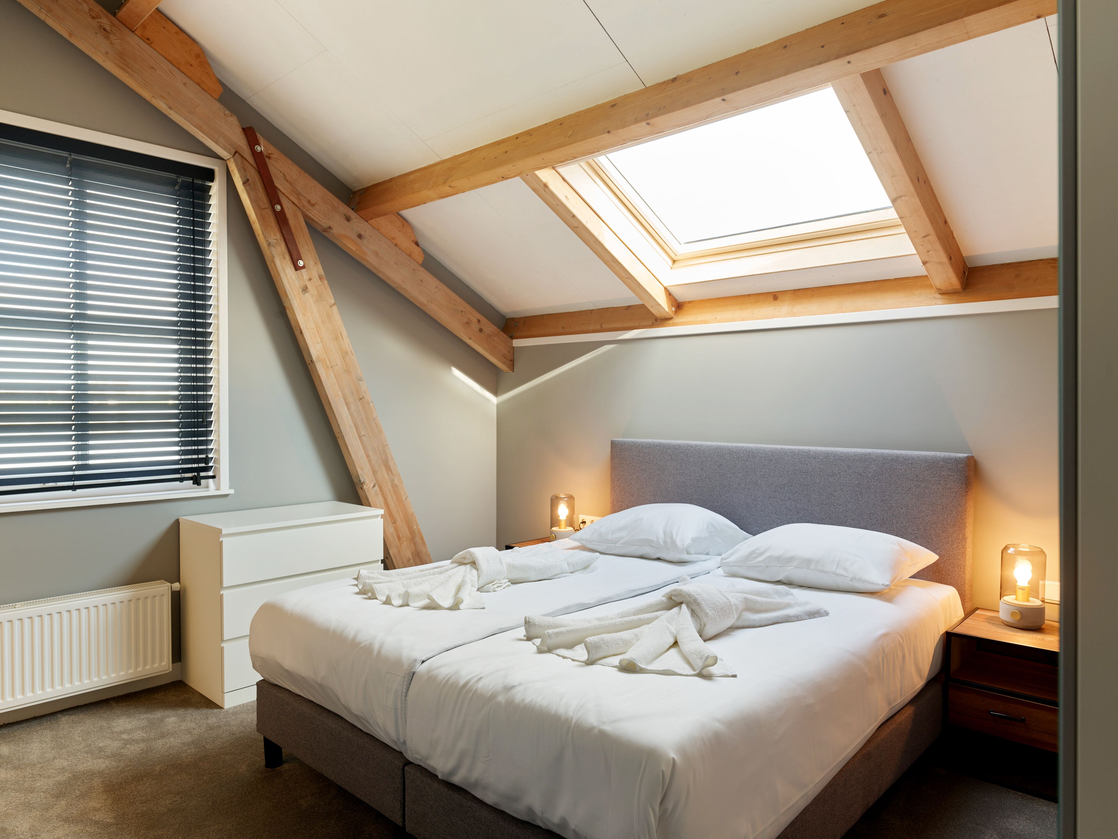 A bedroom with a sloped ceiling, a skylight, a bed with white bedding, a bedside table with a lamp, and a radiator.