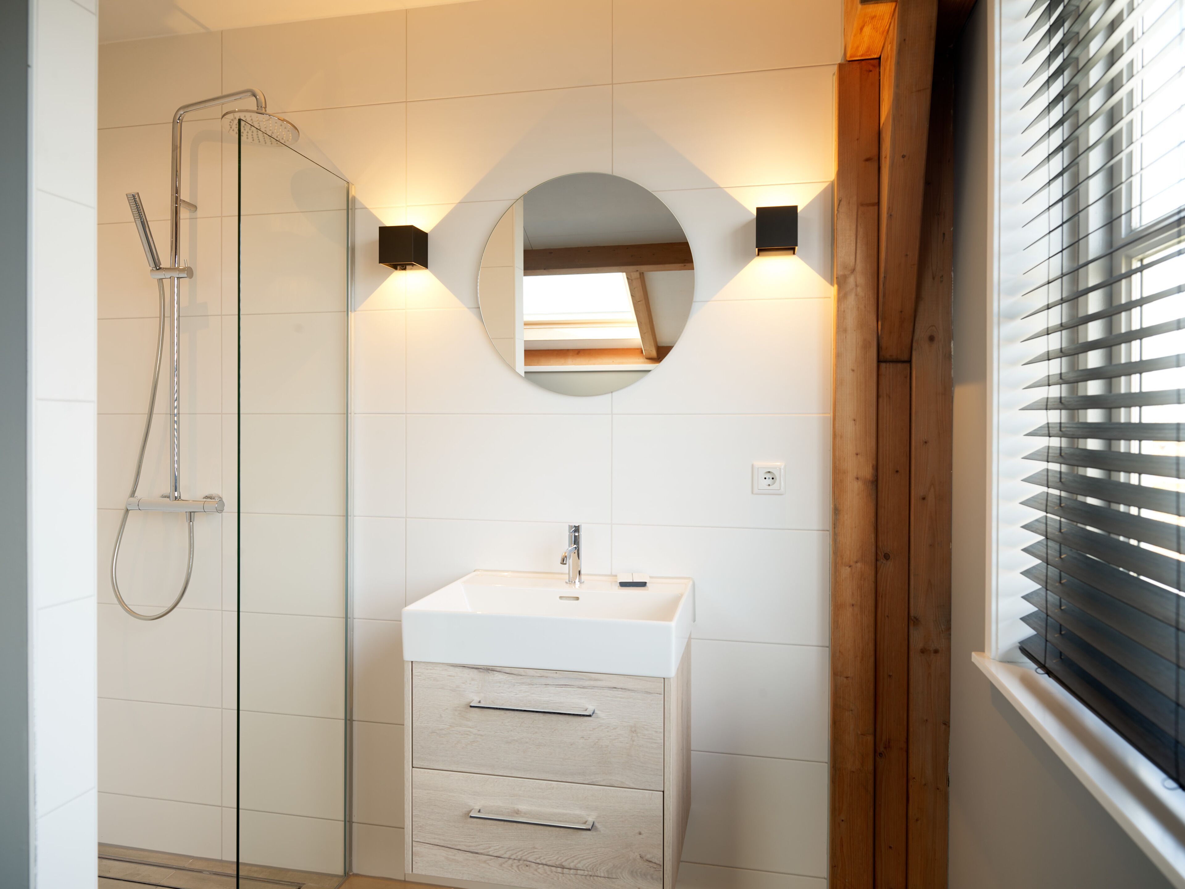 Ein modernes Badezimmer mit Duschkabine aus Glas, einem weißen Waschbecken mit Unterschrank aus Holz und einem runden Spiegel mit Wandleuchten.
