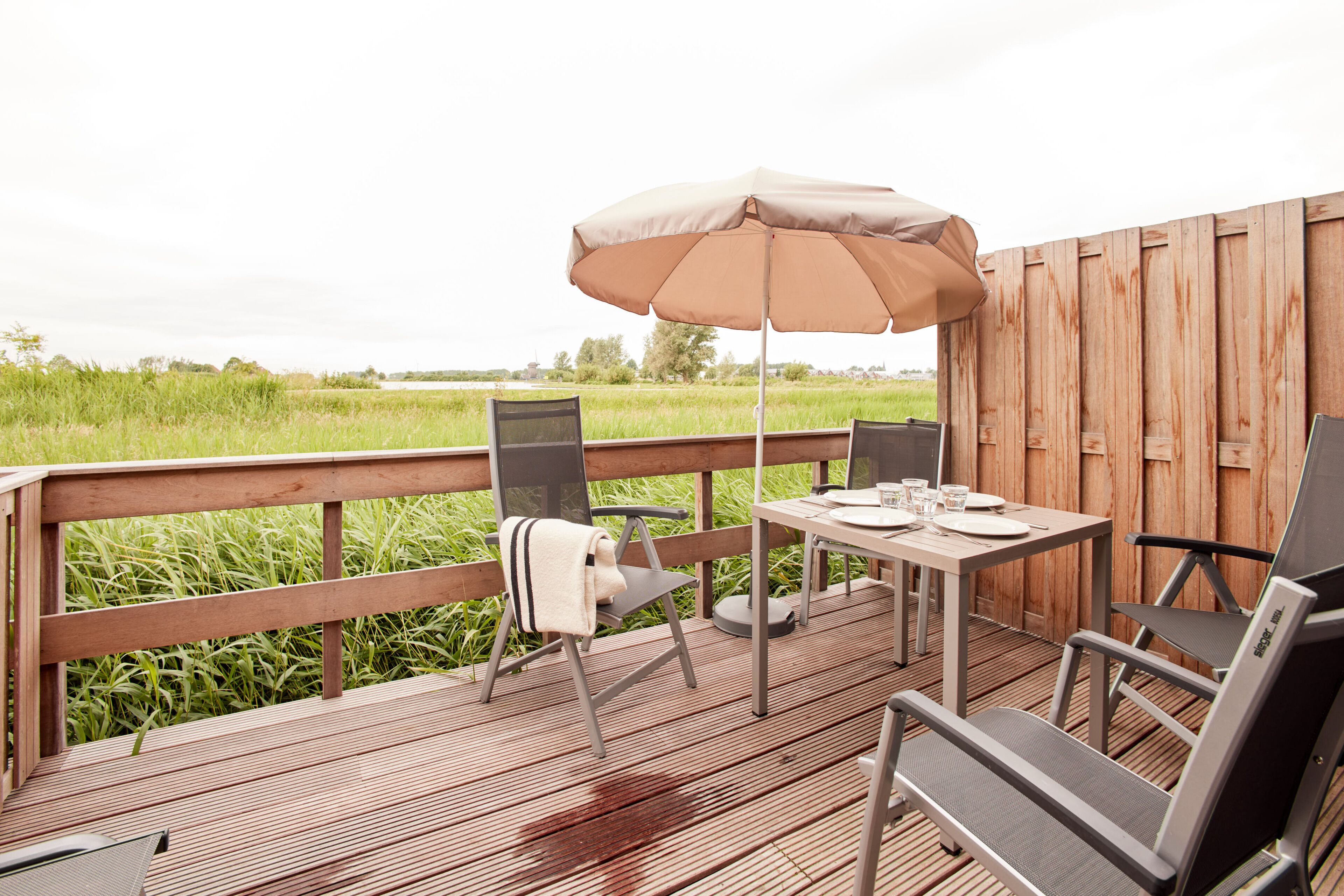 Eine Holzterrasse mit Tisch und Stühlen, einem Sonnenschirm und Blick auf ein grünes Feld.