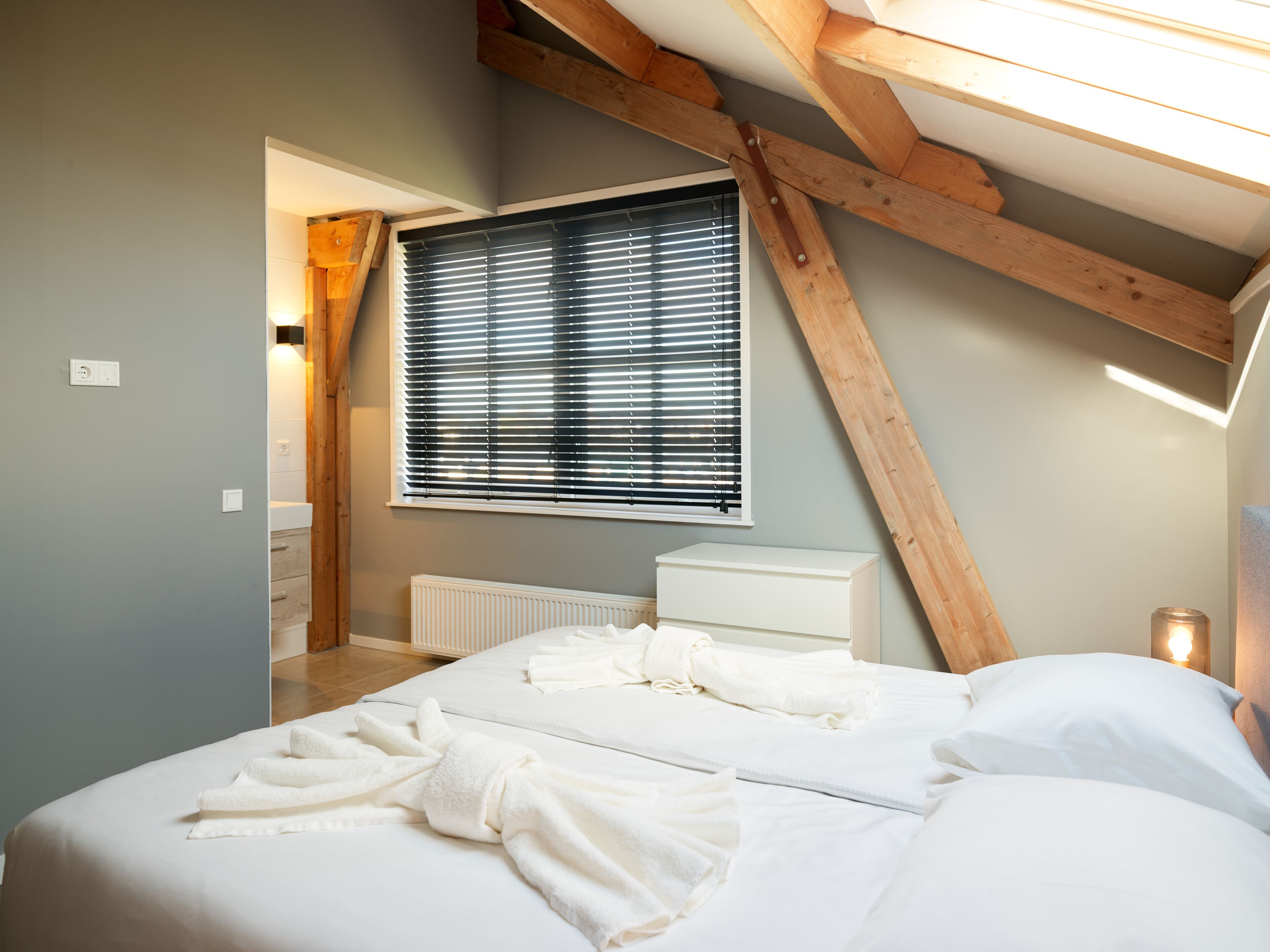 Ein Schlafzimmer mit Schräge, einem Heizkörper, einem Bett mit weißer Bettwäsche und einem Fenster mit Jalousien.