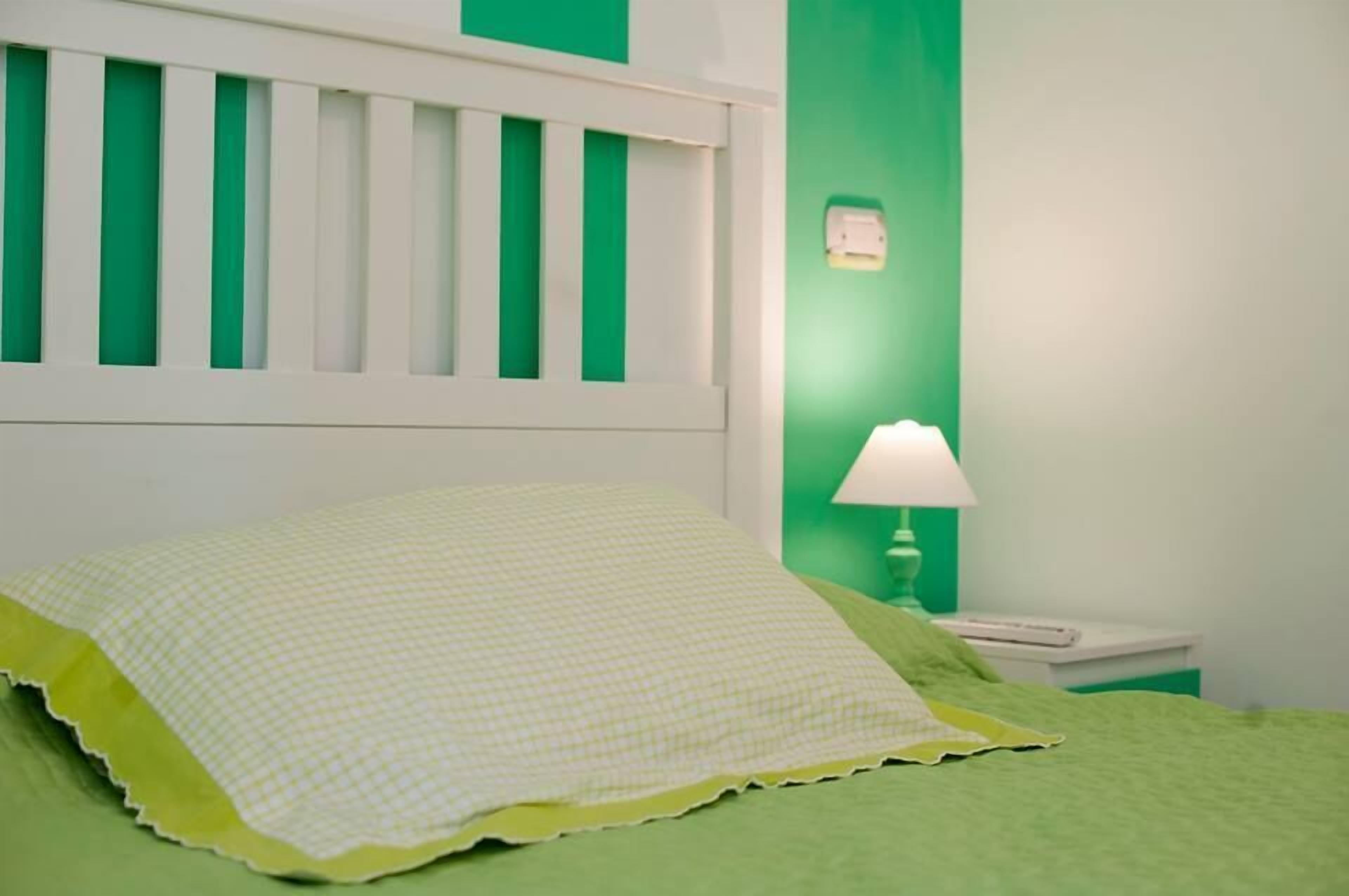 A bed with a green bedspread and a white pillow, a green nightstand with a lamp, and a white headboard with vertical slats.