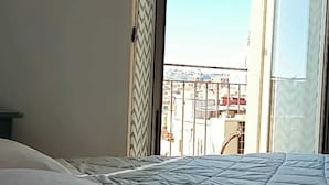 Ein Schlafzimmer mit einem Bett, einem Fenster mit Blick auf Gebäude und einem gemusterten Vorhang.