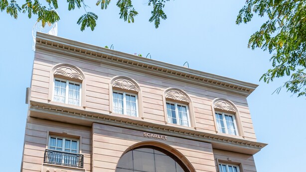 A large, beige building with the name "Scarlet" on the facade, featuring a grand arched entrance, multiple balconies, and a variety of potted plants in front. - Playsales Scarlet (Indore)