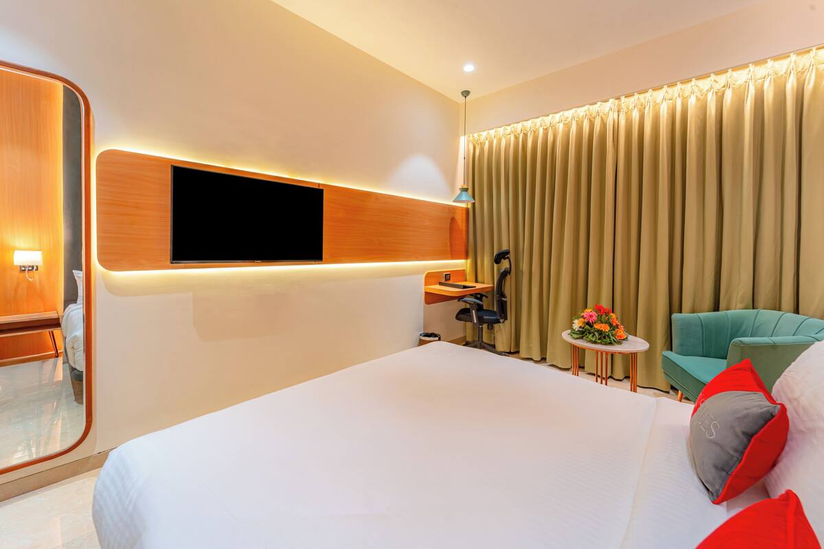 A modern hotel room with a large bed, a flat-screen TV mounted on the wall, a desk with a chair, a small round table with flowers, and a green armchair.