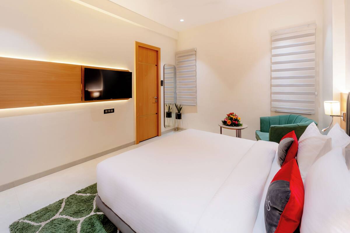 A modern hotel room with a large bed, a flat-screen TV mounted on the wall, a small table with a flower arrangement, and a green armchair.
