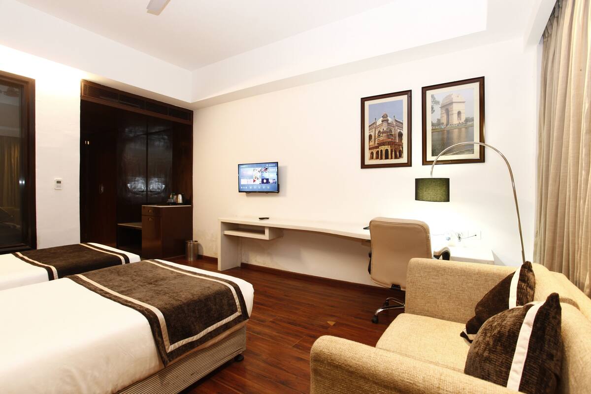 Ein Hotelzimmer mit zwei Betten, einem Schreibtisch, einem Sessel, einem Fernseher und zwei gerahmten Bildern an der Wand.
