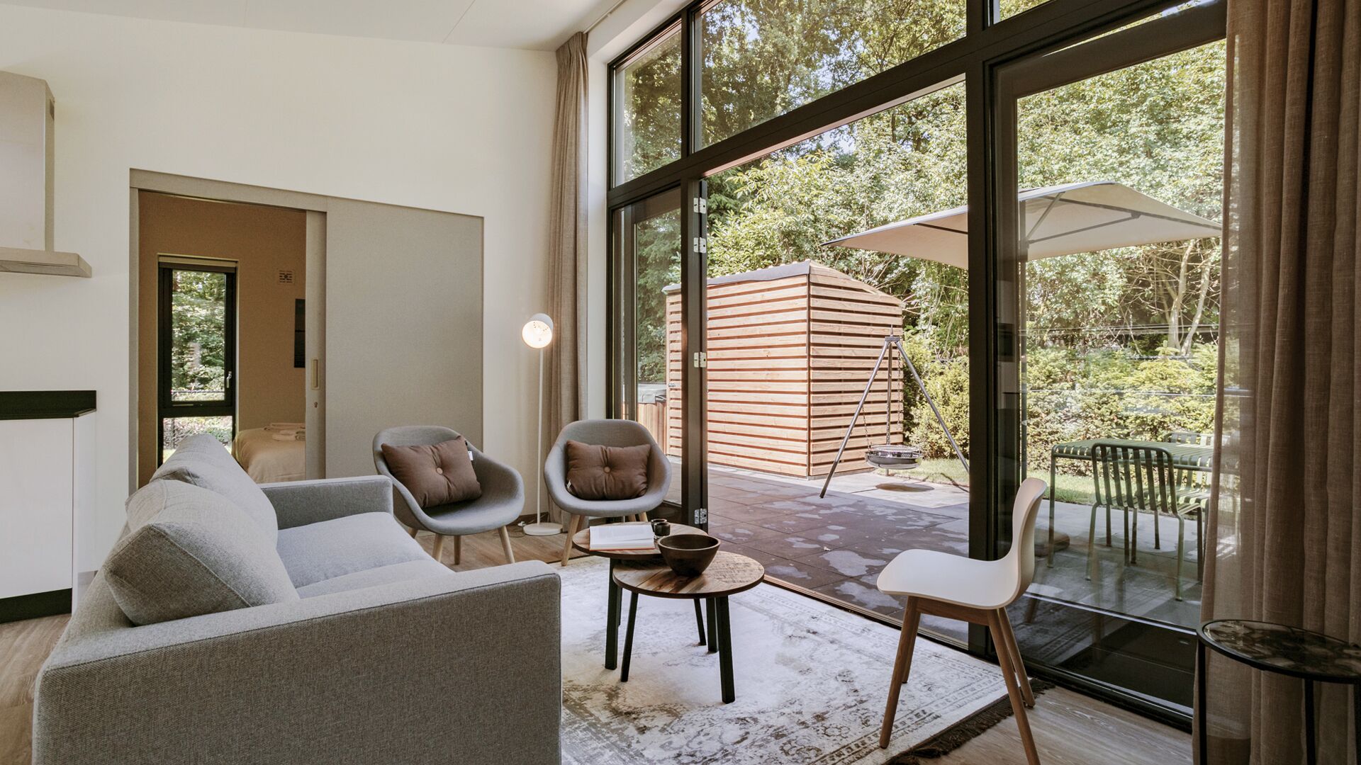 A modern living room with a sofa, two armchairs, a small table, and a large window offering a view of a wooden structure and greenery outside.