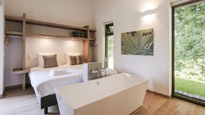 A modern bedroom with a large bathtub, a bed with patterned pillows, and a view of greenery through a window. - Suitelodges Gooilanden (Loosdrecht)