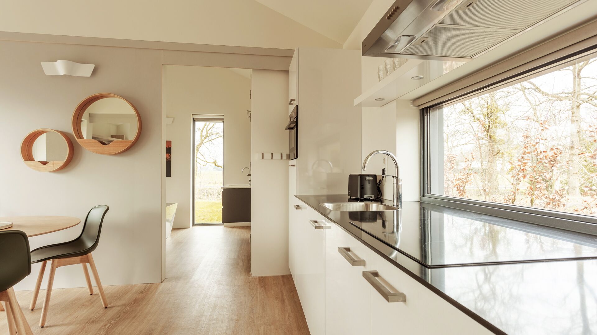 A modern kitchen with a large window, a sink, and a kettle. There is a dining area with a table and chairs.