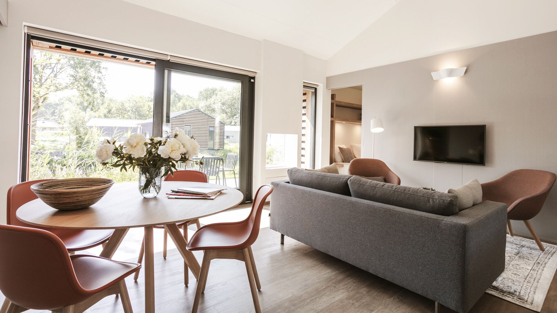 A modern living room with a grey sofa, wooden chairs, a dining table with a vase of flowers, and a TV mounted on the wall.