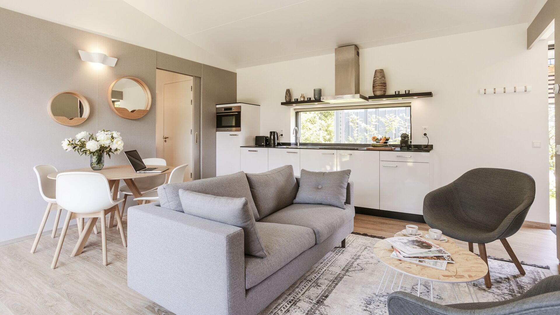 Een moderne woonkamer met een grijze bank, een salontafel en een grijze fauteuil. Er is een zithoek met een tafel en stoelen, en een keuken met witte kastjes en een raam.
