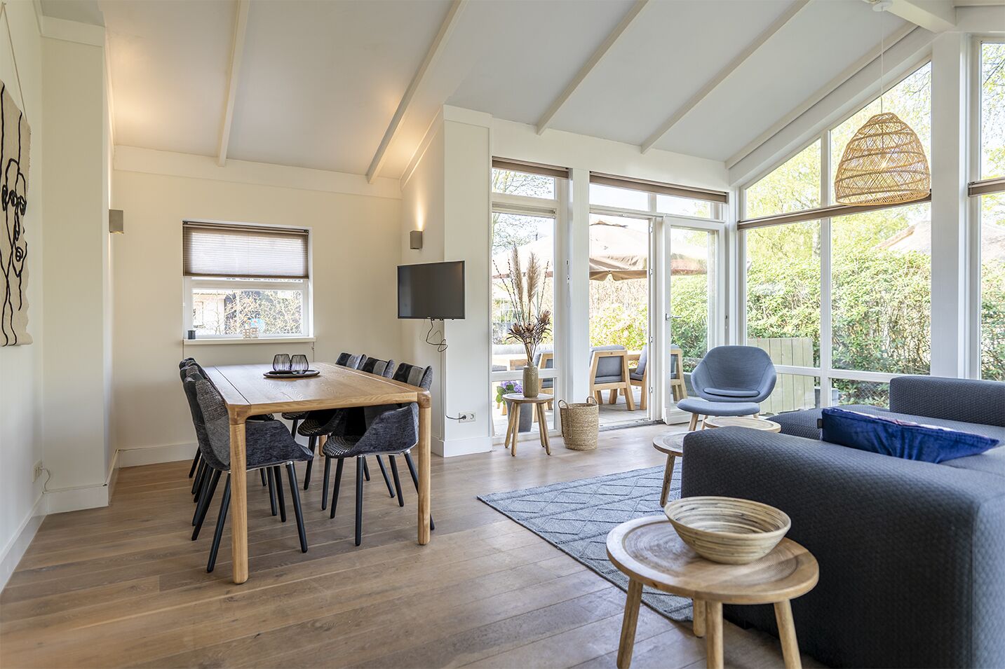 A modern living room with a wooden dining table, grey upholstered chairs, a grey sofa, and a large window offering a view of greenery.