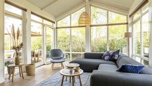 A spacious room with a large window, a grey sofa, a round wooden table, and a wicker basket. - Suitelodges Gooilanden (Loosdrecht)