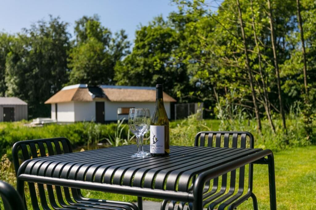 Een metalen tafel met een fles wijn en een glas, omringd door stoelen, buiten geplaatst met begroeiing en een gebouw op de achtergrond.