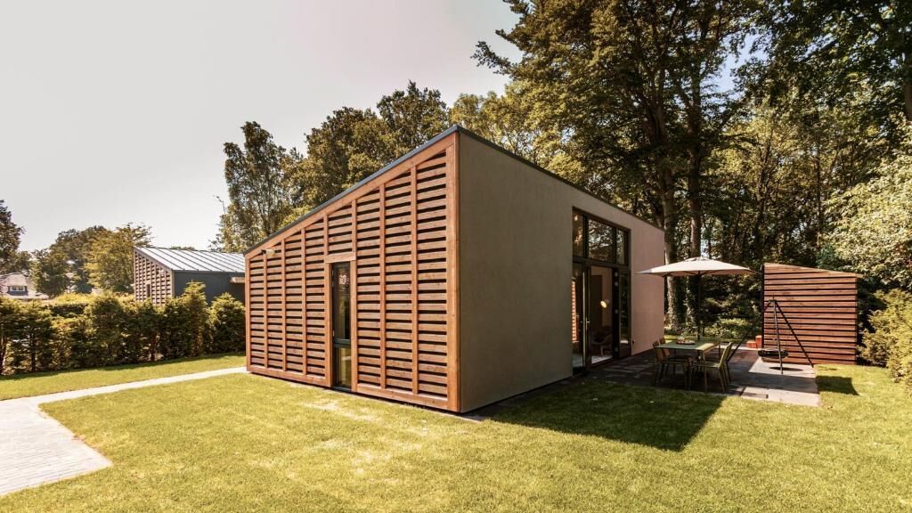 Un edificio moderno y rectangular con fachada de listones, una puerta visible y un pequeño espacio al aire libre con una mesa y sillas.