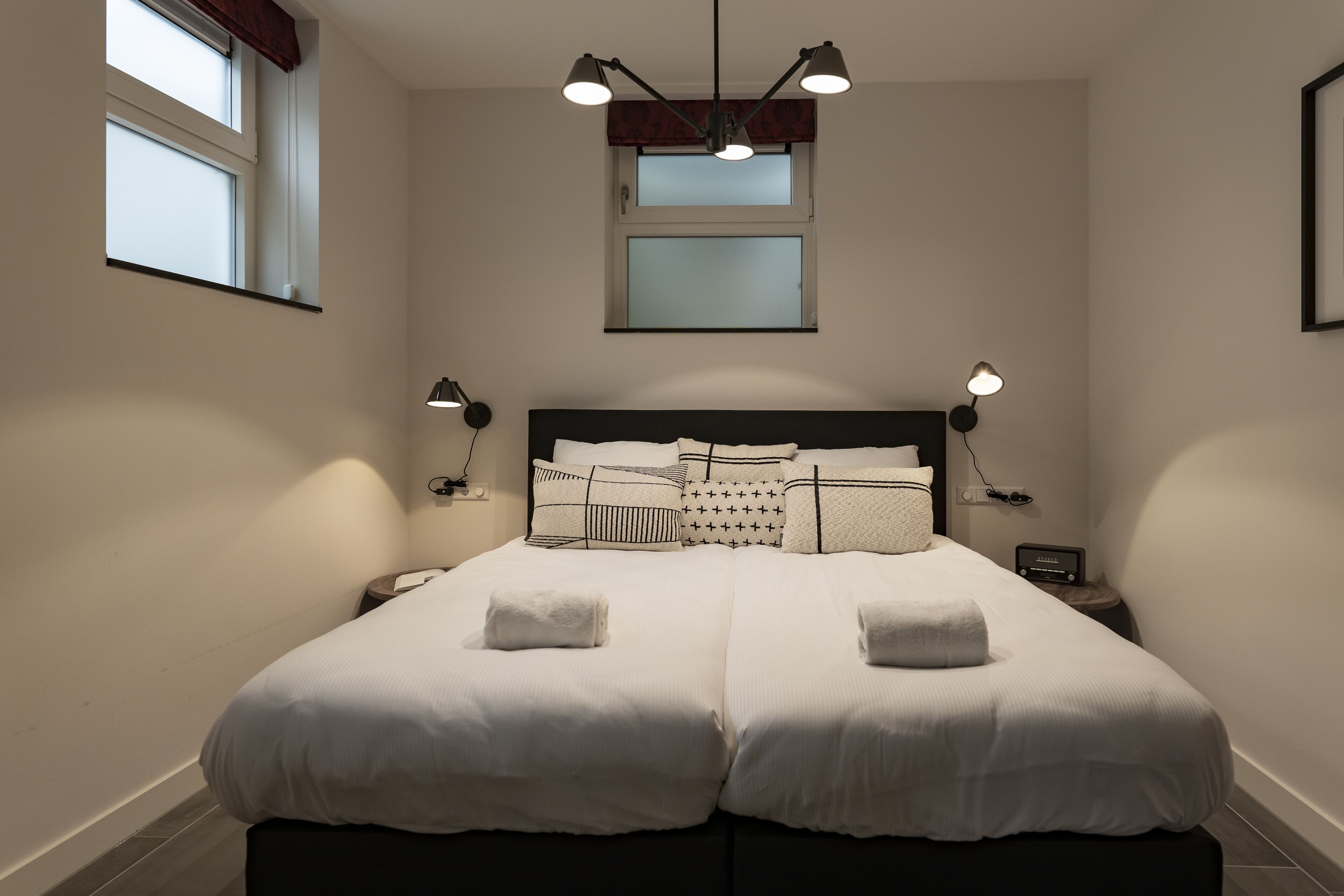 Ein Doppelbett mit weißer Bettwäsche, zwei Nachttische mit Lampen, eine Deckenleuchte und ein Fenster mit Jalousien.
