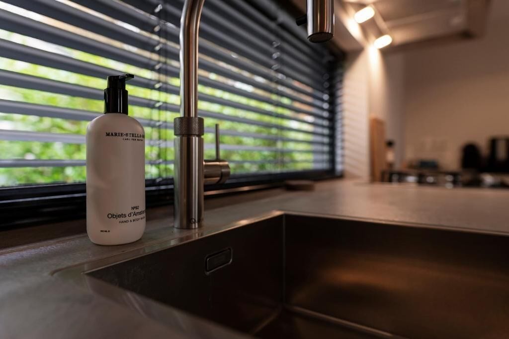 A bathroom with a modern faucet, a bottle of Marie-Stella hand and body wash, and a window with blinds.
