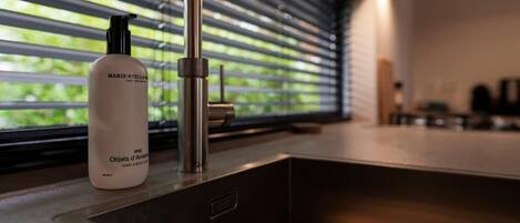 A bathroom with a modern faucet, a bottle of Marie-Stella hand and body wash, and a window with blinds.