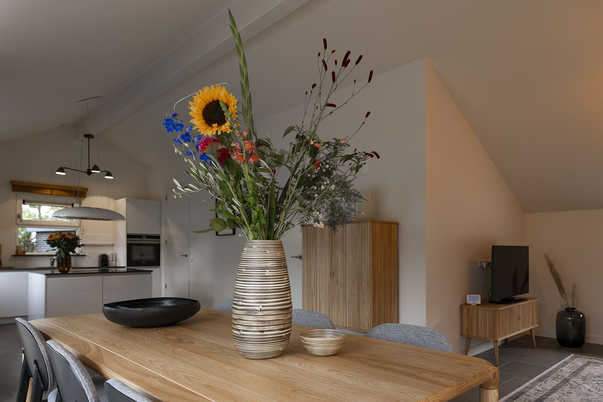 A dining table with a striped vase containing a sunflower and assorted flowers, a black bowl, and a small bowl.