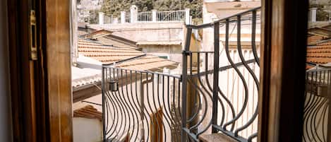 A balcony with a wrought iron railing overlooking a residential area with terracotta roofs and a hillside in the background.