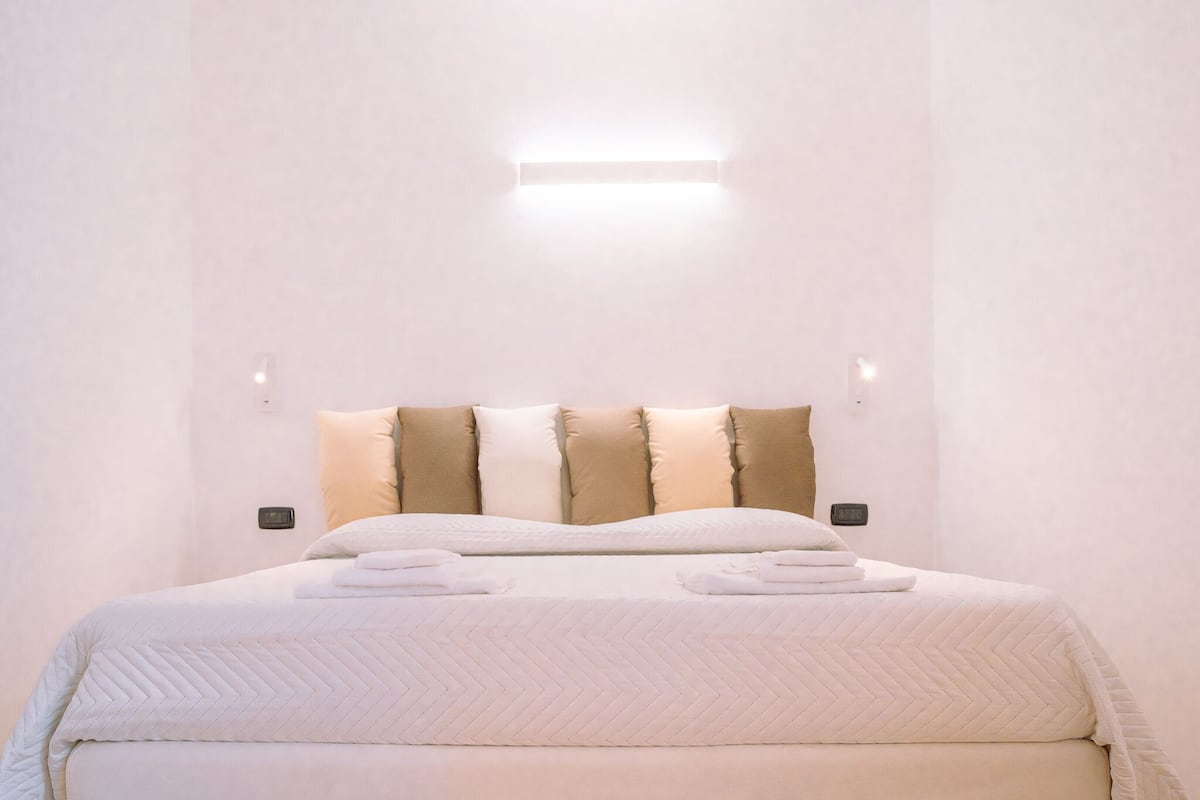 A neatly made bed with a white quilt, multiple pillows, and a bedside table with a lamp.