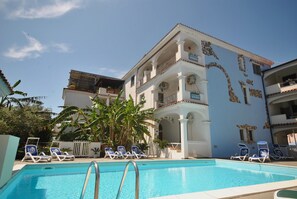 A swimming pool with lounge chairs, a building with balconies, and a clear sky. - 3.5-room apartment (70m²) on first floor with veranda measuring 10m²  (Sardegna)