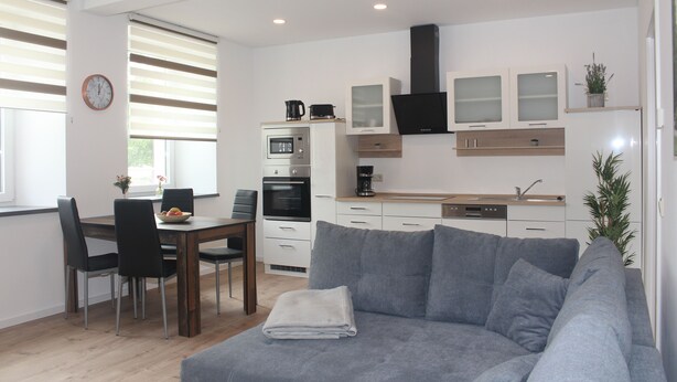 Una sala de estar moderna con un sofá gris, una mesa de comedor con sillas, y una zona de cocina con armarios y electrodomésticos.