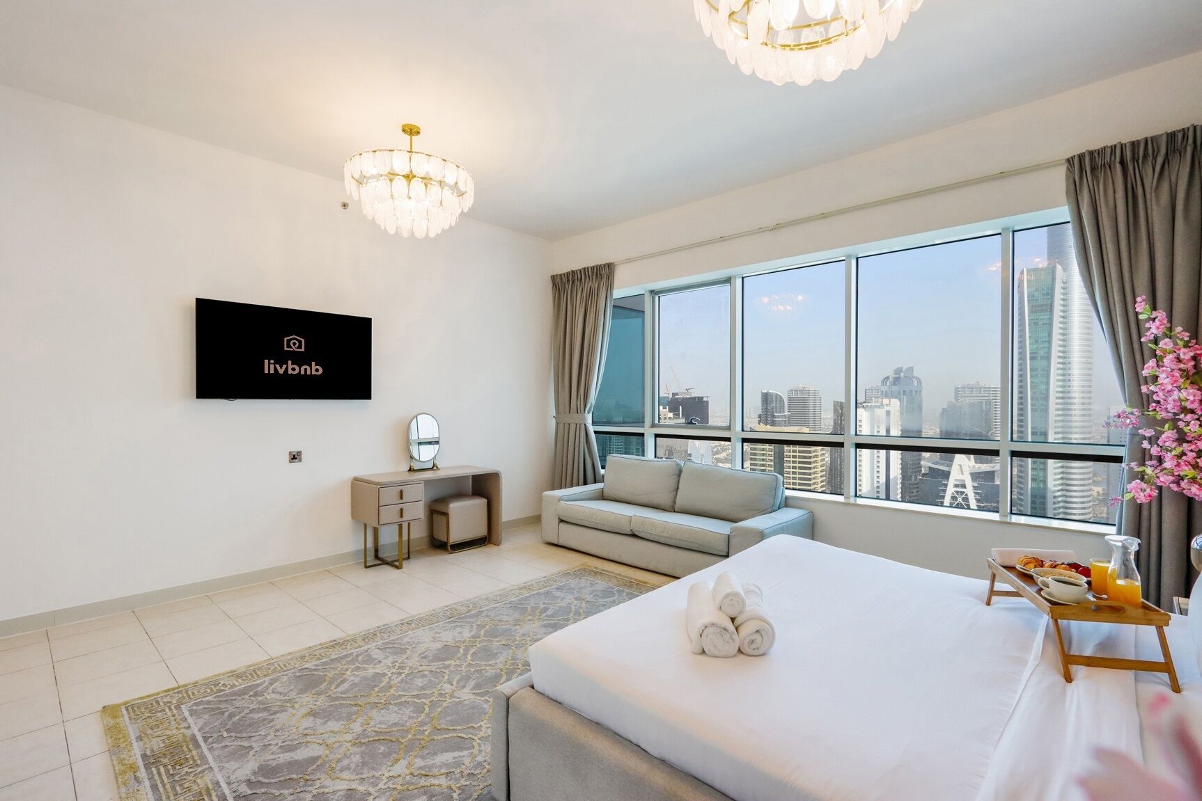 A modern hotel room with a large bed, a desk with a mirror, a TV mounted on the wall, and a view of the city skyline through large windows.