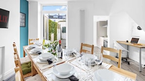 A dining area with a long table set for six, a view of the outside, and a kitchen in the background.