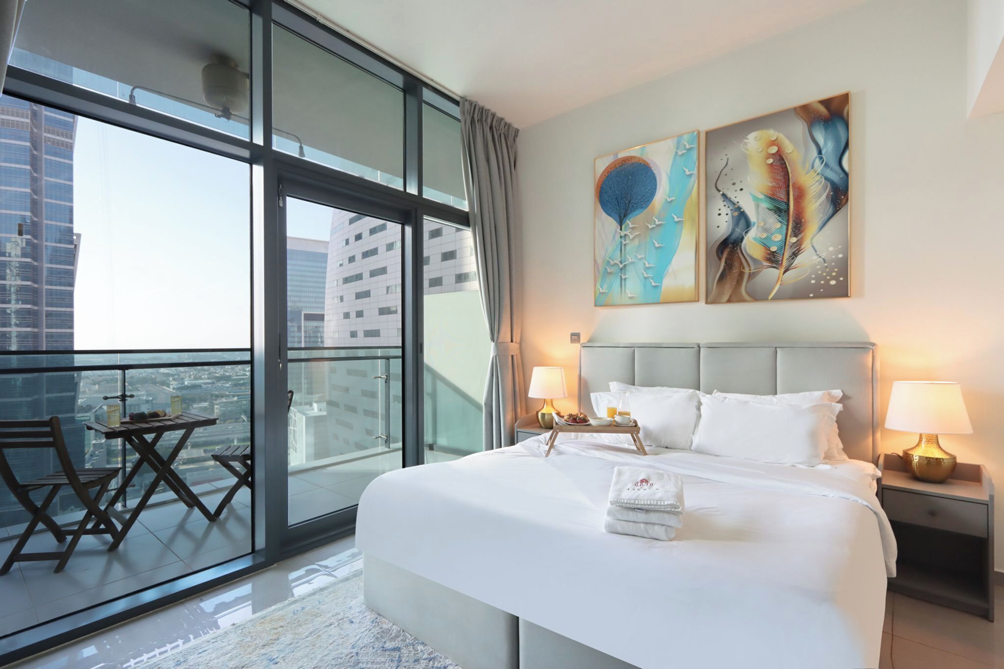 A hotel room with a large bed, two bedside lamps, a view of the city through floor-to-ceiling windows, and a balcony with a table and chairs.